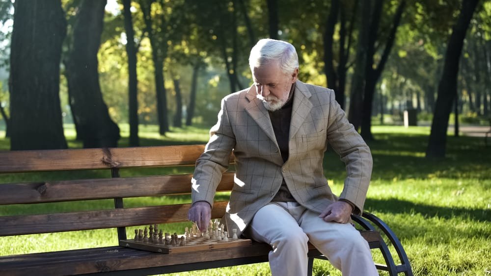 Old man sitting on bench in park alone and playing chess