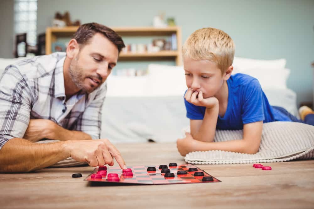 father and son playing checkers game while lying on floor